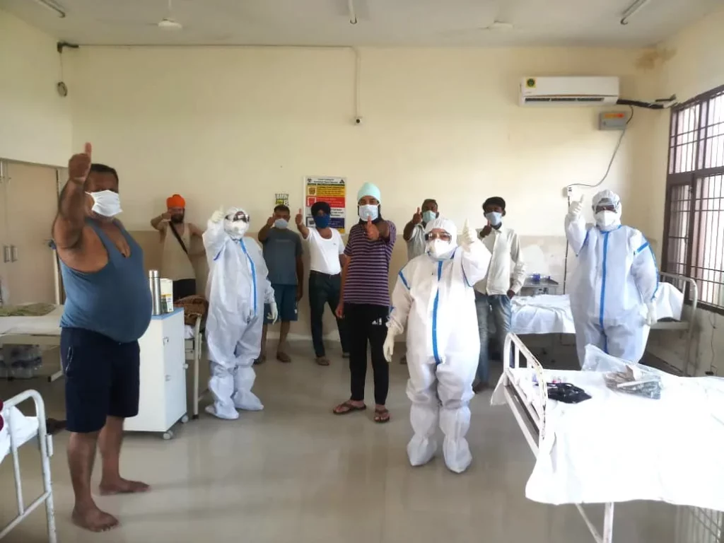 Patients and healthcare workers in PPE stand in a ward at a hospital in Punjab and make a thumbs up sign towards the camera. 