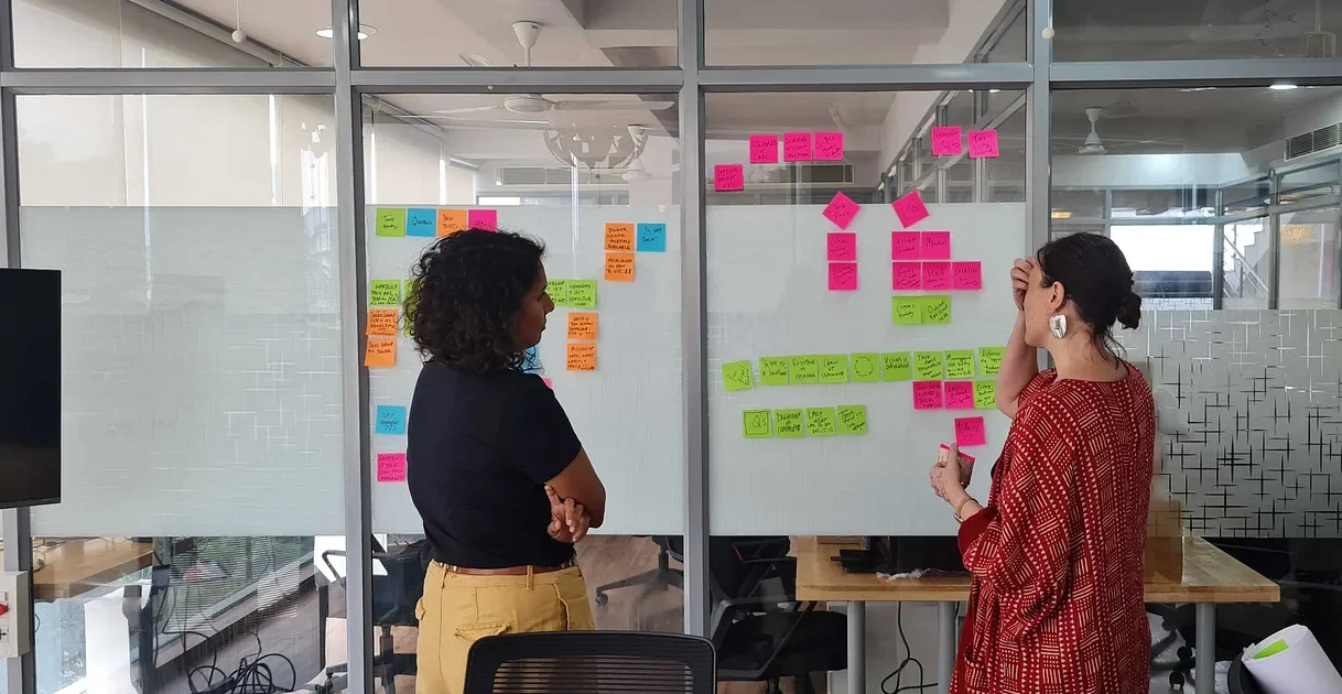 Two members of the Caregiving Lab at a brainstorm session in the Noora Health office. They are standing in front of a glass wall with colorful sticky notes and with their backs to the camera.