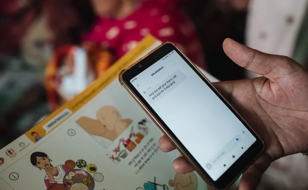 A close up of a hand holding a mobile phone with a text message and Noora Health training materials in the background.