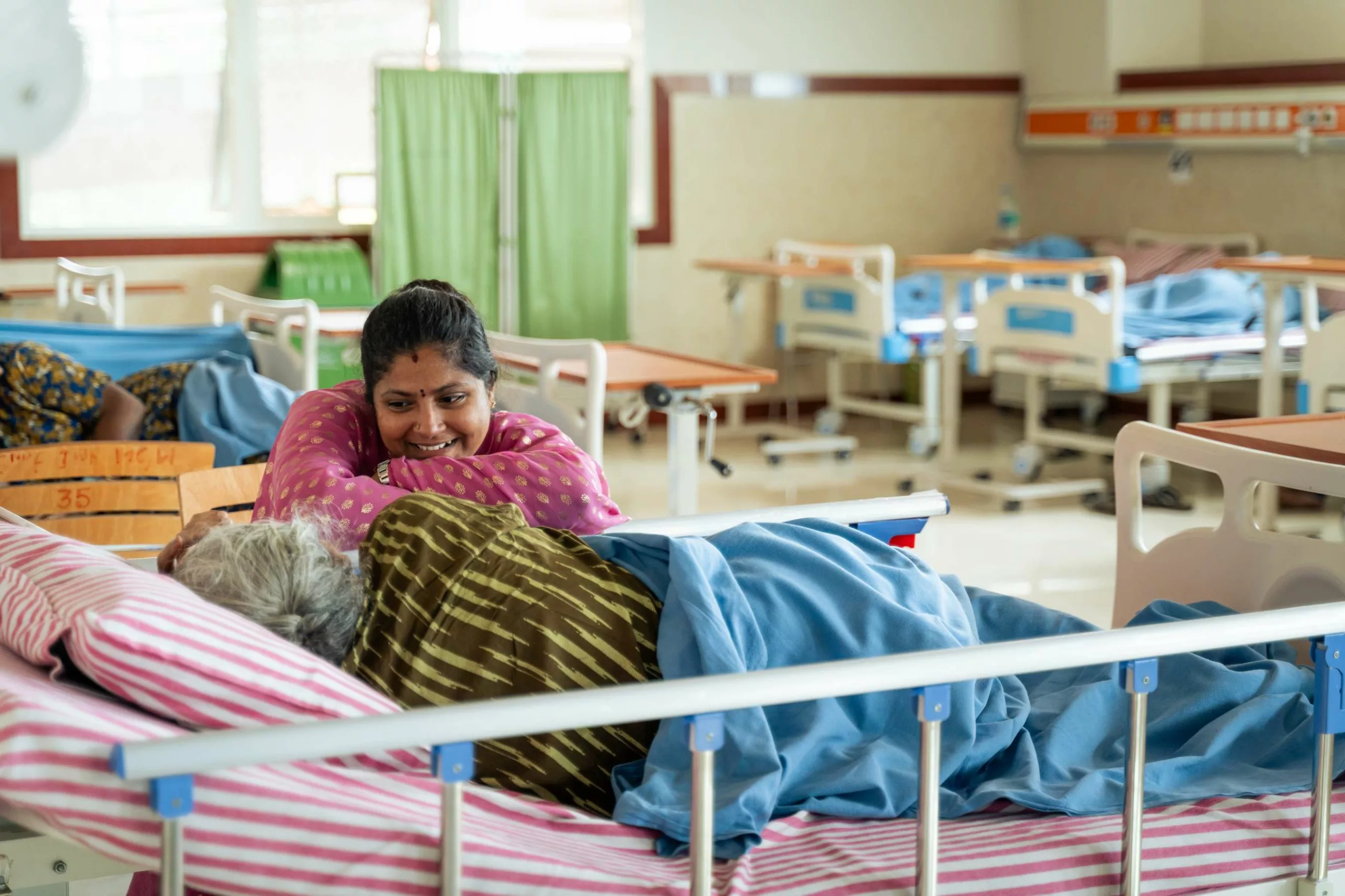 A smiling caregiver leans against a patient's bed at the Sri Jayadeva Institute of Cardiovascular Sciences and Research, in Bangalore, India.